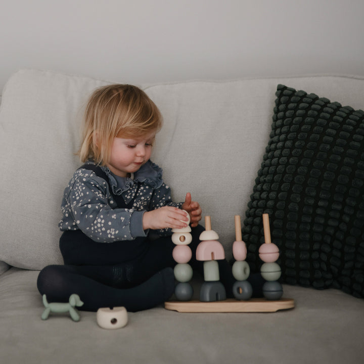 A toddler sitting on a bed playing with a wooden multi-shape stacker toy by Mushie & Co.