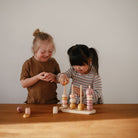 Two young girls sitting side-by-side at a table, playing together with a wooden multi-shape stacker toy by Mushie & Co.
