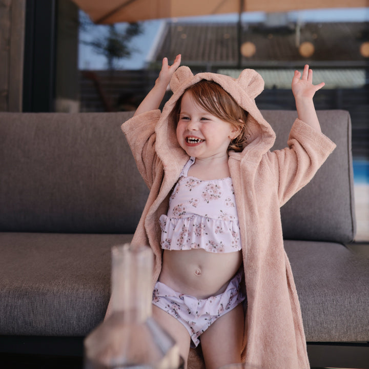 Child wearing a floral swimsuit and pink robe with bear ears, standing on a couch.