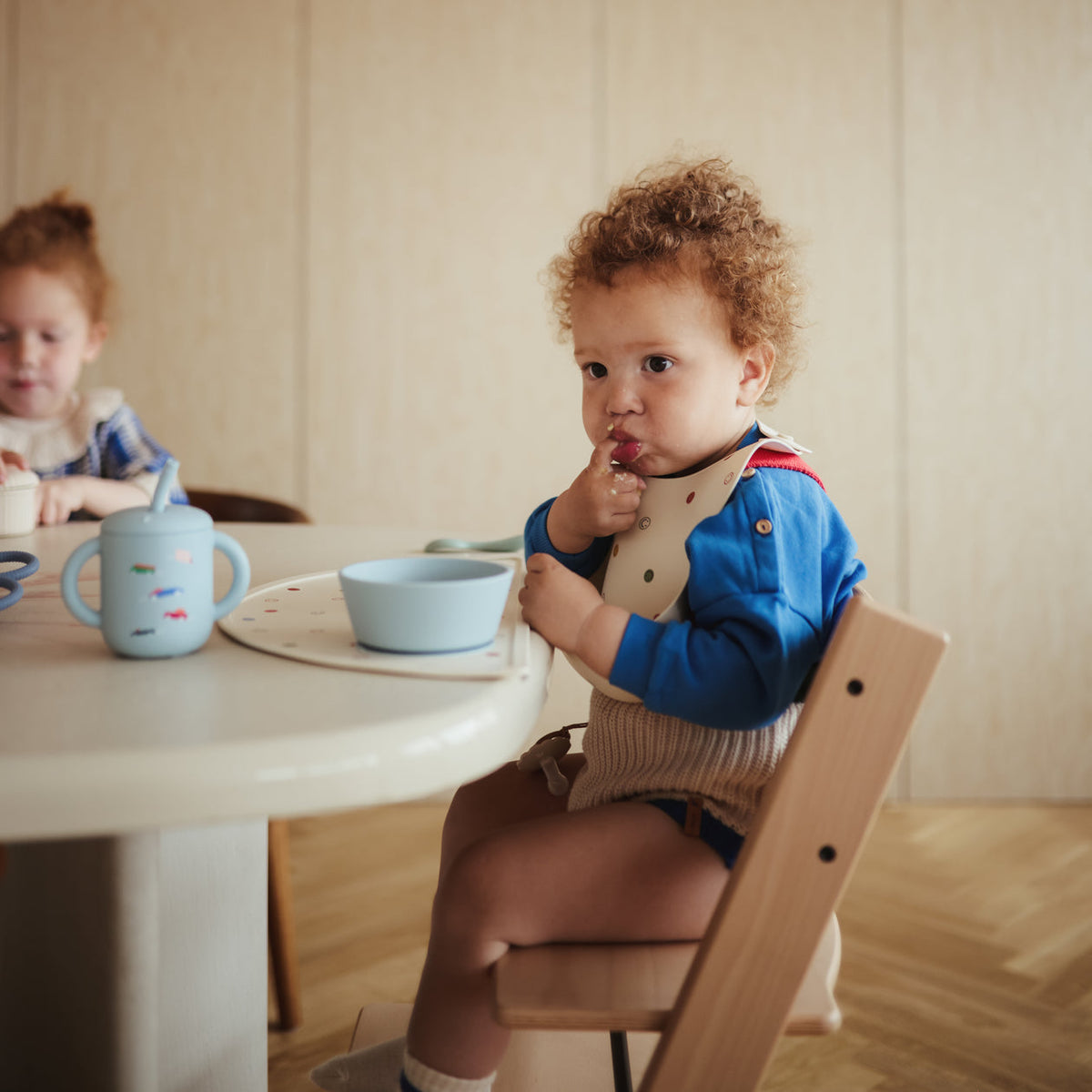 Two children sitting at a table with blue cups and bowls, one child is eating.