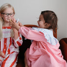 Two young girls sitting together, one in a red and white striped dress and the other in a pink outfit, sharing an object.