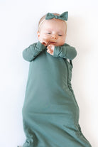 A baby wearing a muted sage green color long-sleeve wrap swaddle and a matching bow headband, lying on a plain white background.