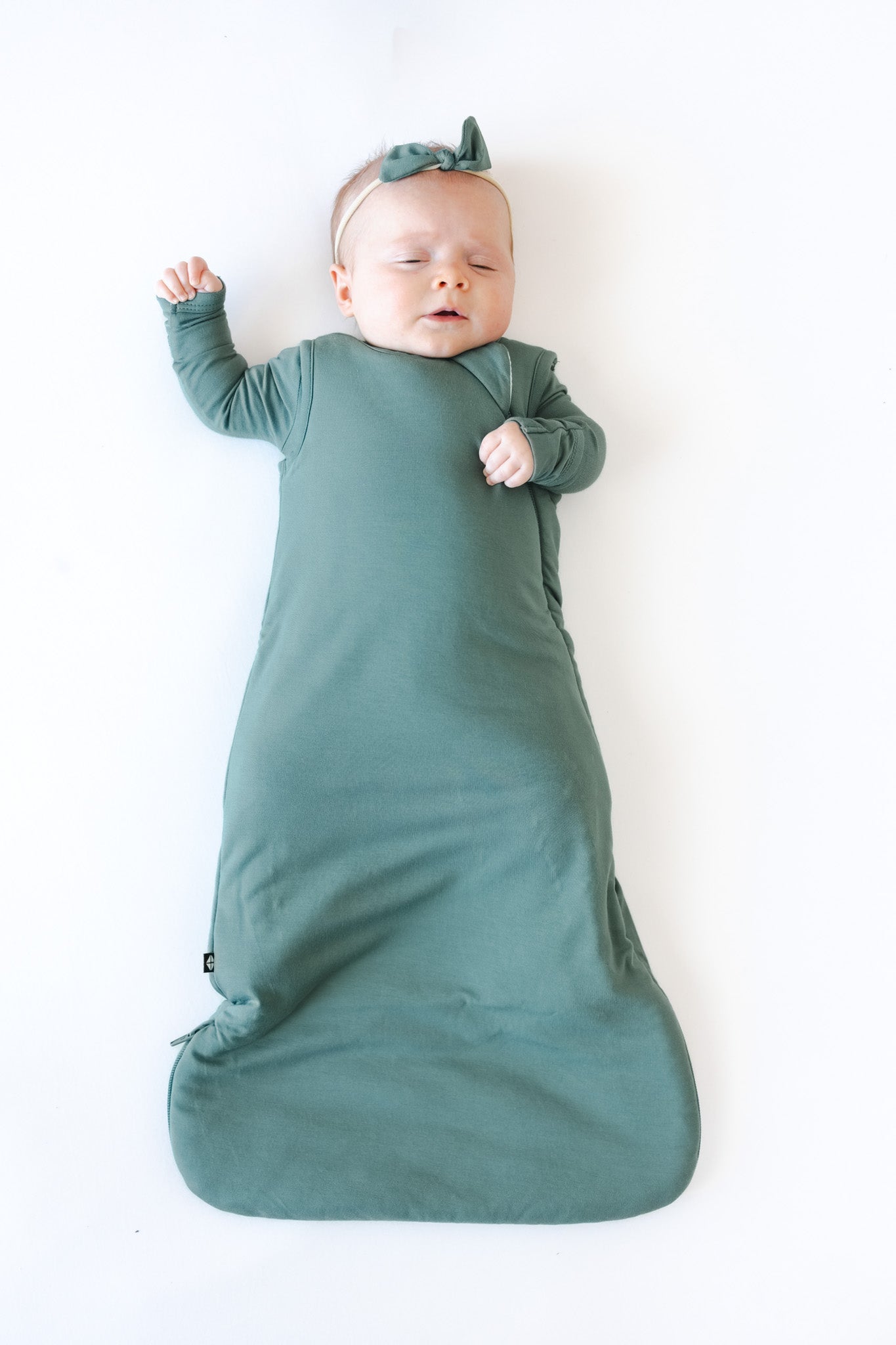 A baby lying on their back wearing a muted sage green color long-sleeve sleep sack and matching bow headband, set against a plain white background.