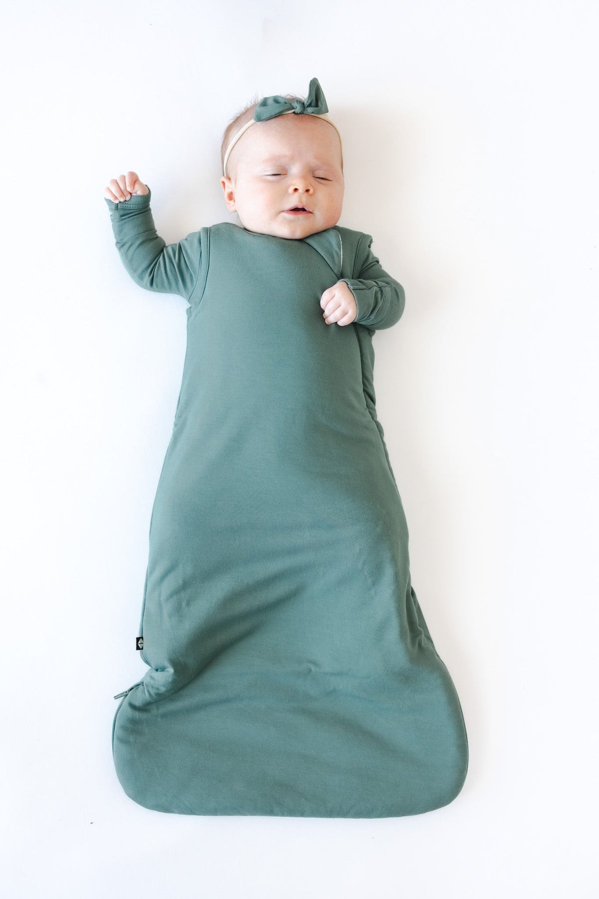 A baby lying on their back wearing a muted sage green color long-sleeve sleep sack and matching bow headband, set against a plain white background.