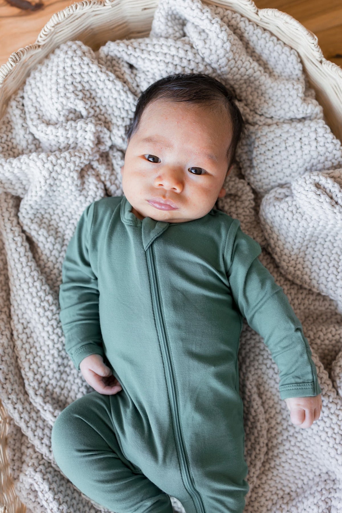 A newborn baby wearing a sage green long-sleeve zip-up footless onesie, lying on a chunky knit cream blanket inside a woven basket.