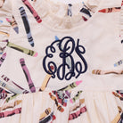 Close-up of a cream-colored children's dress with a colorful crayon print and a navy blue monogram embroidered on the chest.