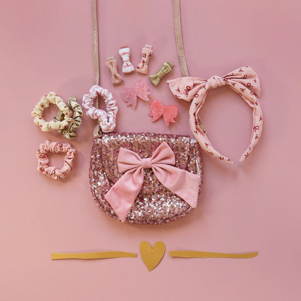 Collection of pink and gold hair accessories including a purse, headband, and clips on a pink background.