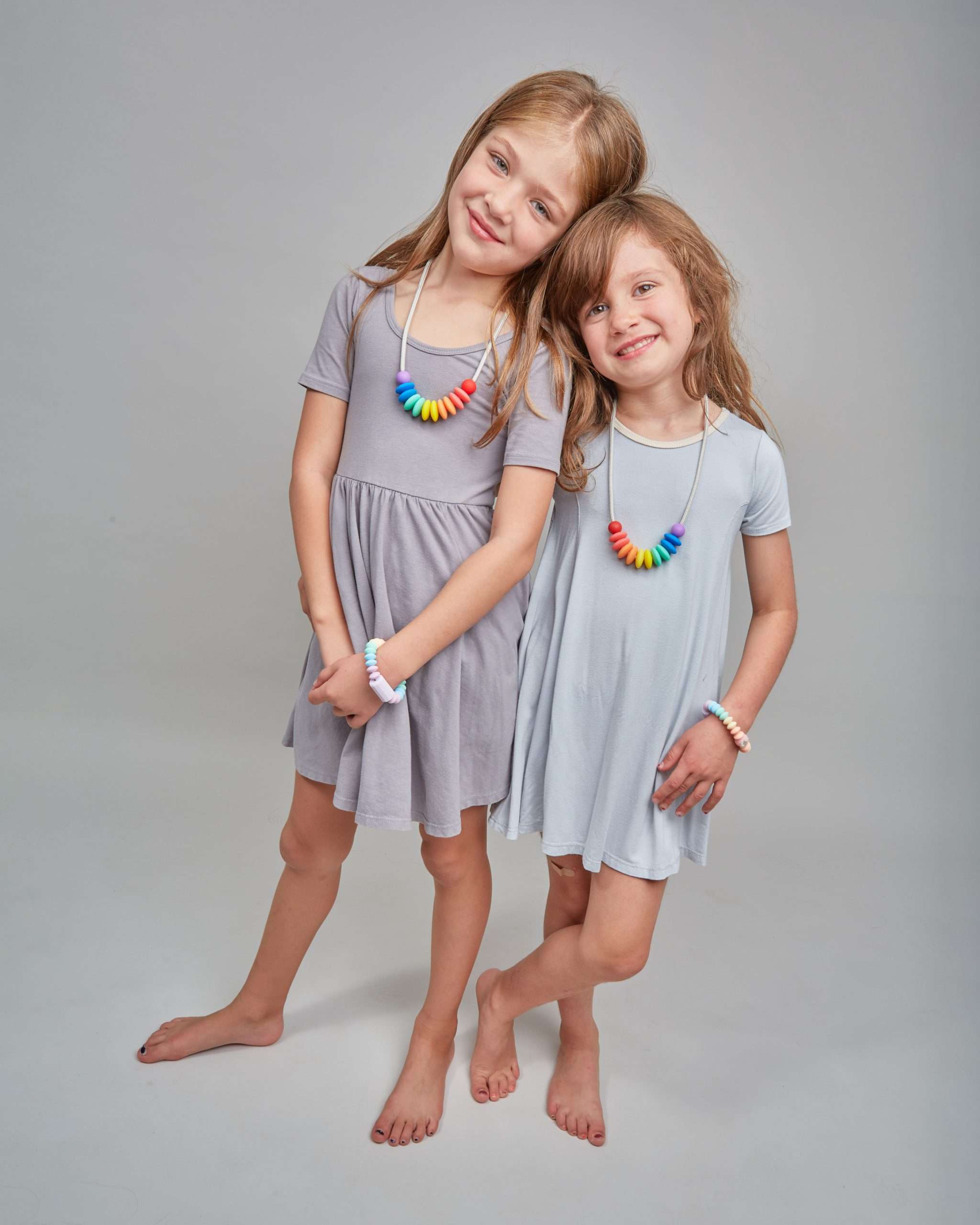 Two young girls wearing gray dresses with colorful necklaces on a plain background
