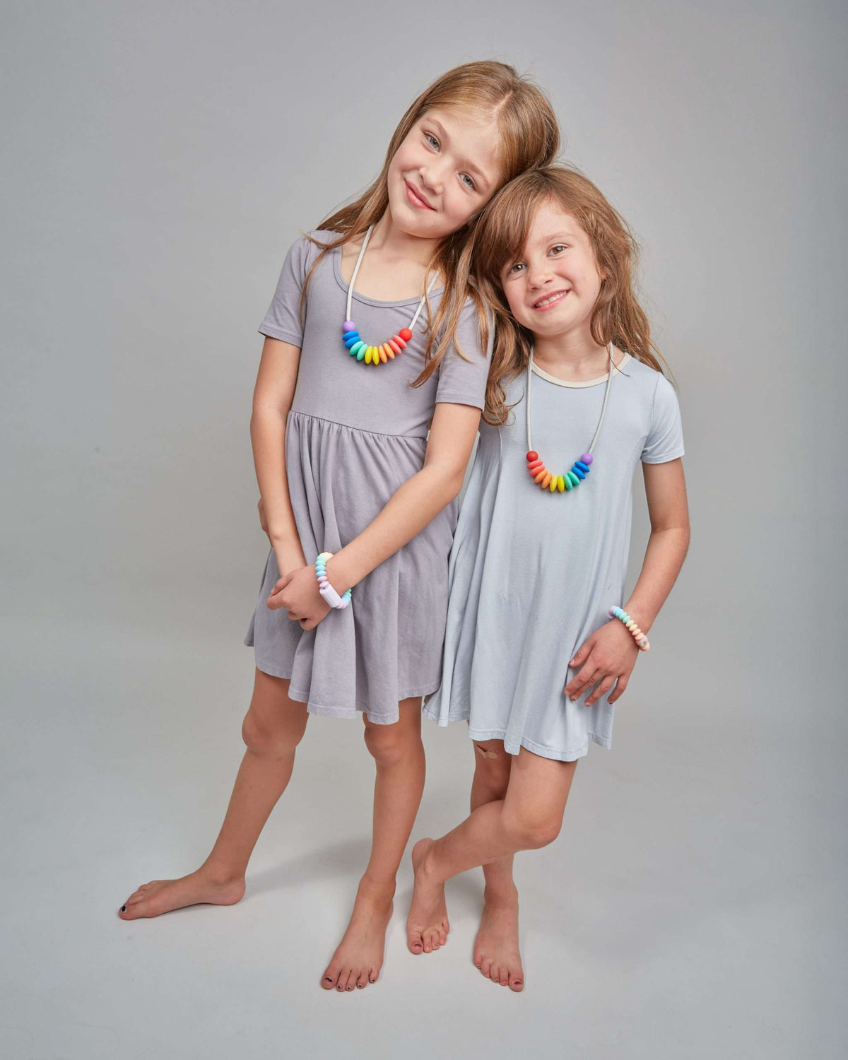 Two young girls wearing gray dresses with colorful necklaces on a plain background