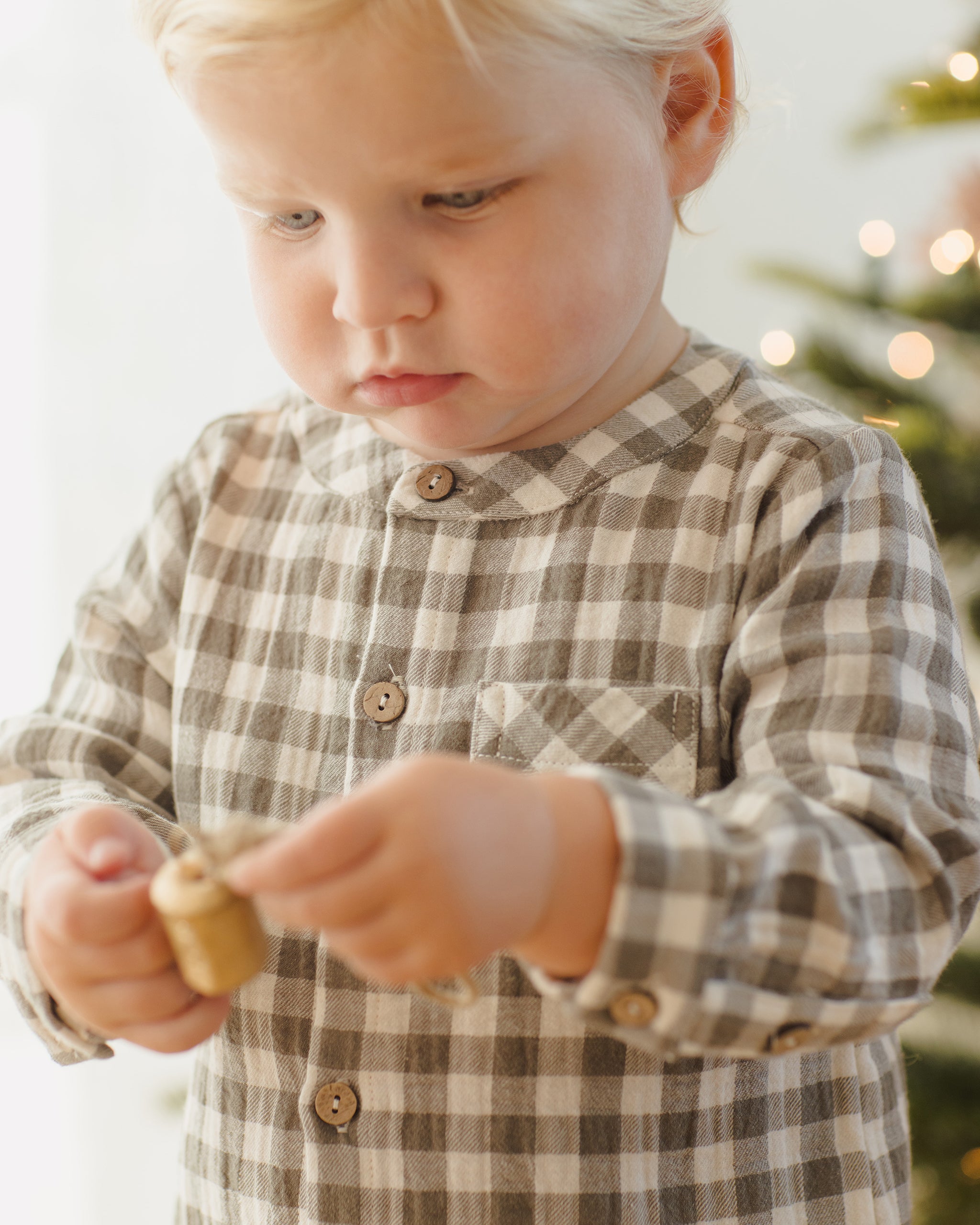 Child wearing a plaid shirt holding a small gold ornament with a blurred Christmas tree in the background.