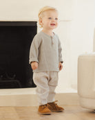 Child wearing a gray shirt and checkered pants standing in a room with a fireplace.