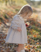 A young girl with blonde hair wearing a light blue and white checkered dress, standing outdoors in a wooded area with fallen leaves and holding a red apple.