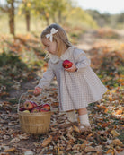 A young girl in a beige and white checkered dress with a white bow in her hair bends down to a wooden basket filled with apples on a leaf-covered path in an autumn park.