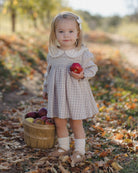 A young girl stands outdoors in a leaf-covered path holding a red apple, wearing a long-sleeve plaid dress with a white Peter Pan collar. A small wooden basket filled with apples sits beside her.