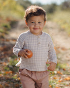 A toddler boy wearing a beige and white checkered long-sleeve henley shirt and dusty pink pants, holding an orange in an outdoor setting.