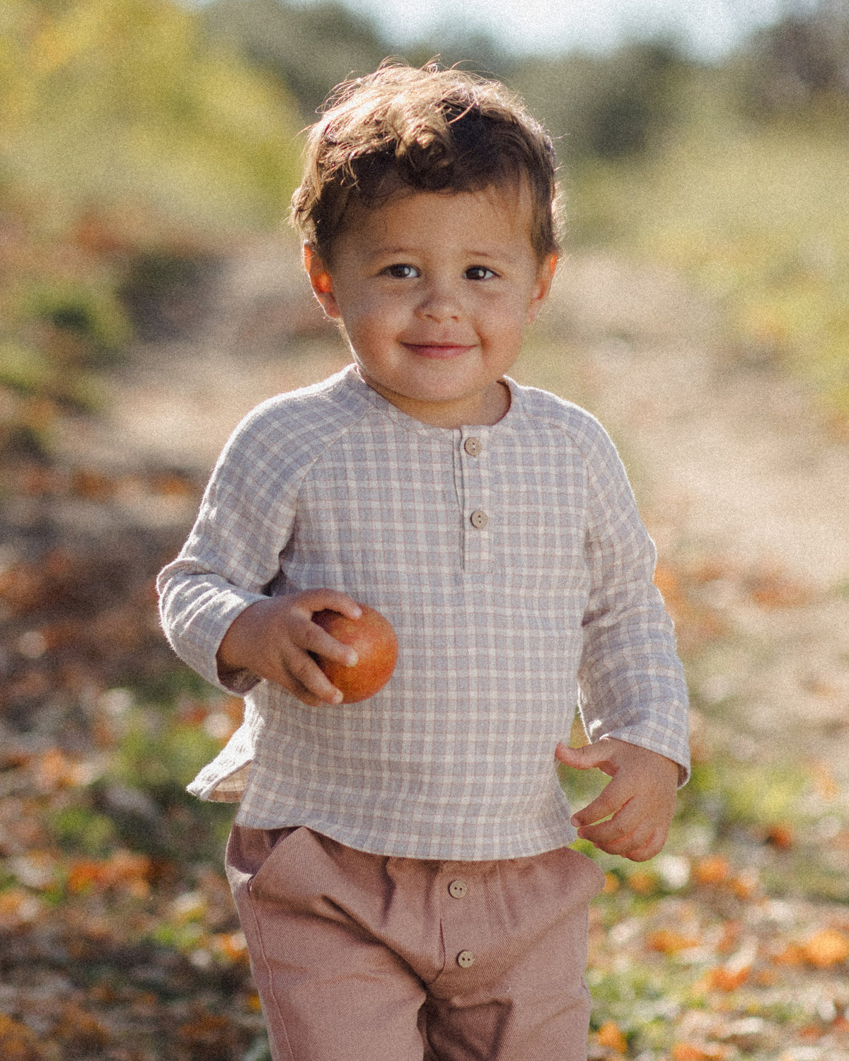 A toddler boy wearing a beige and white checkered long-sleeve henley shirt and dusty pink pants, holding an orange in an outdoor setting.