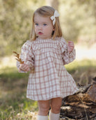 A young toddler girl wearing a pink and white gingham dress with a white bow in her hair, standing outdoors in a grassy, wooded area.