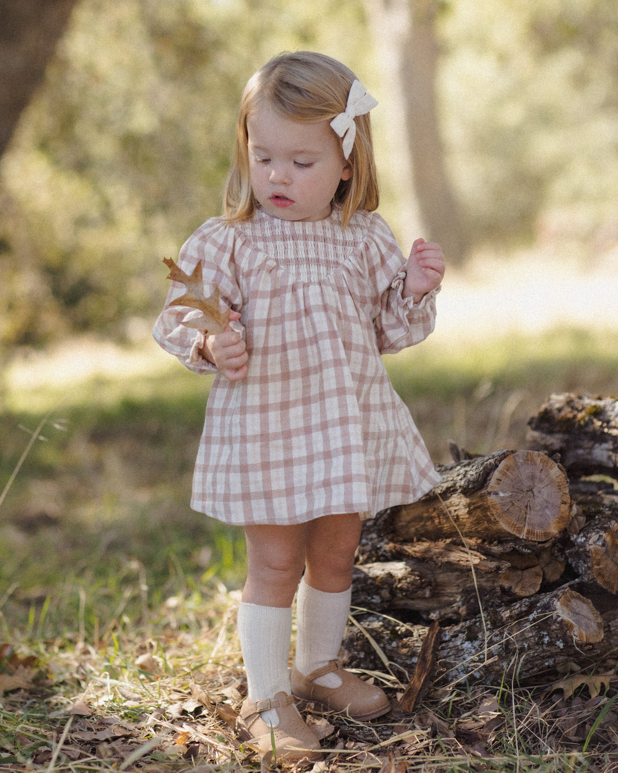 A young girl stands outdoors in a wooded setting wearing a pink and white gingham long-sleeve dress, cream knee-high socks, and tan shoes. She has a white bow in her hair and is standing near a log pile.