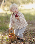 A toddler girl in a beige and white checkered shirt and light gray pants stands outdoors in a grassy, sunlit area, holding a red apple and a small woven basket.