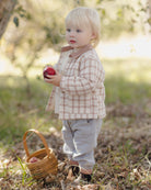 A toddler standing outdoors in a wooded setting, wearing a beige and white gingham button-up shirt and light gray pants, holding a red apple next to a small woven basket.
