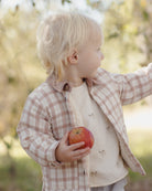 A toddler with blonde hair wearing a beige and brown plaid button-up jacket over a cream fleece shirt, holding a red apple in an outdoor garden setting.