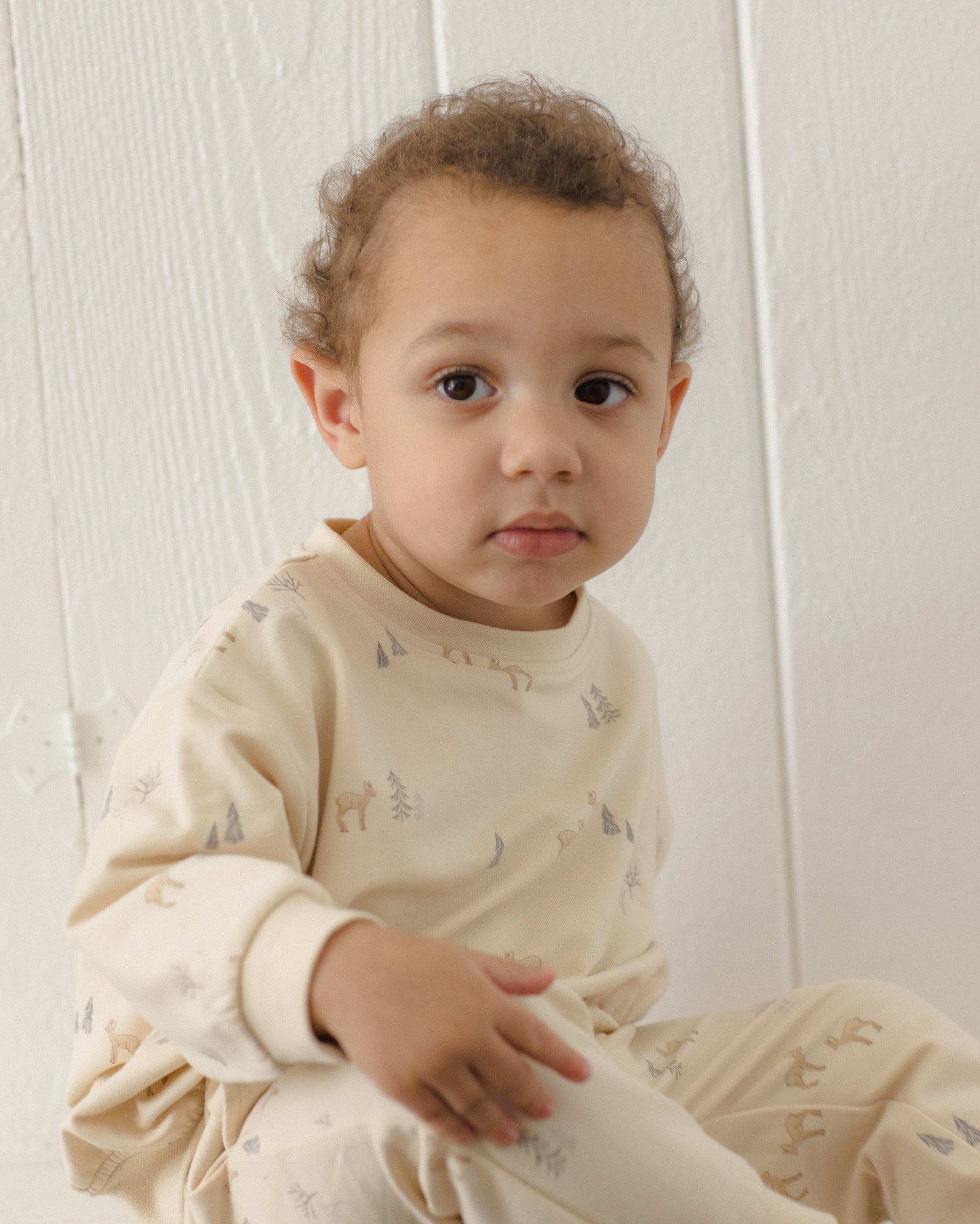 A toddler wearing a cream-colored long-sleeve pajama set with a small gray animal print, sitting against a white paneled wall.