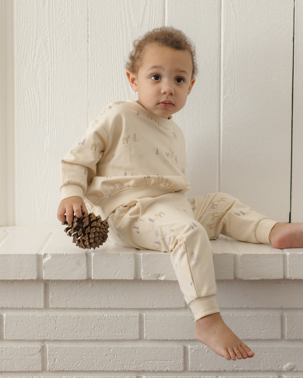 A toddler wearing a cream-colored pajama set with a subtle pine tree pattern sits on a white brick ledge holding a pinecone against a white paneled wall.