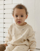 A toddler wearing a cream-colored long-sleeve pajama set with a subtle deer and tree print, sitting in front of a white paneled wall.