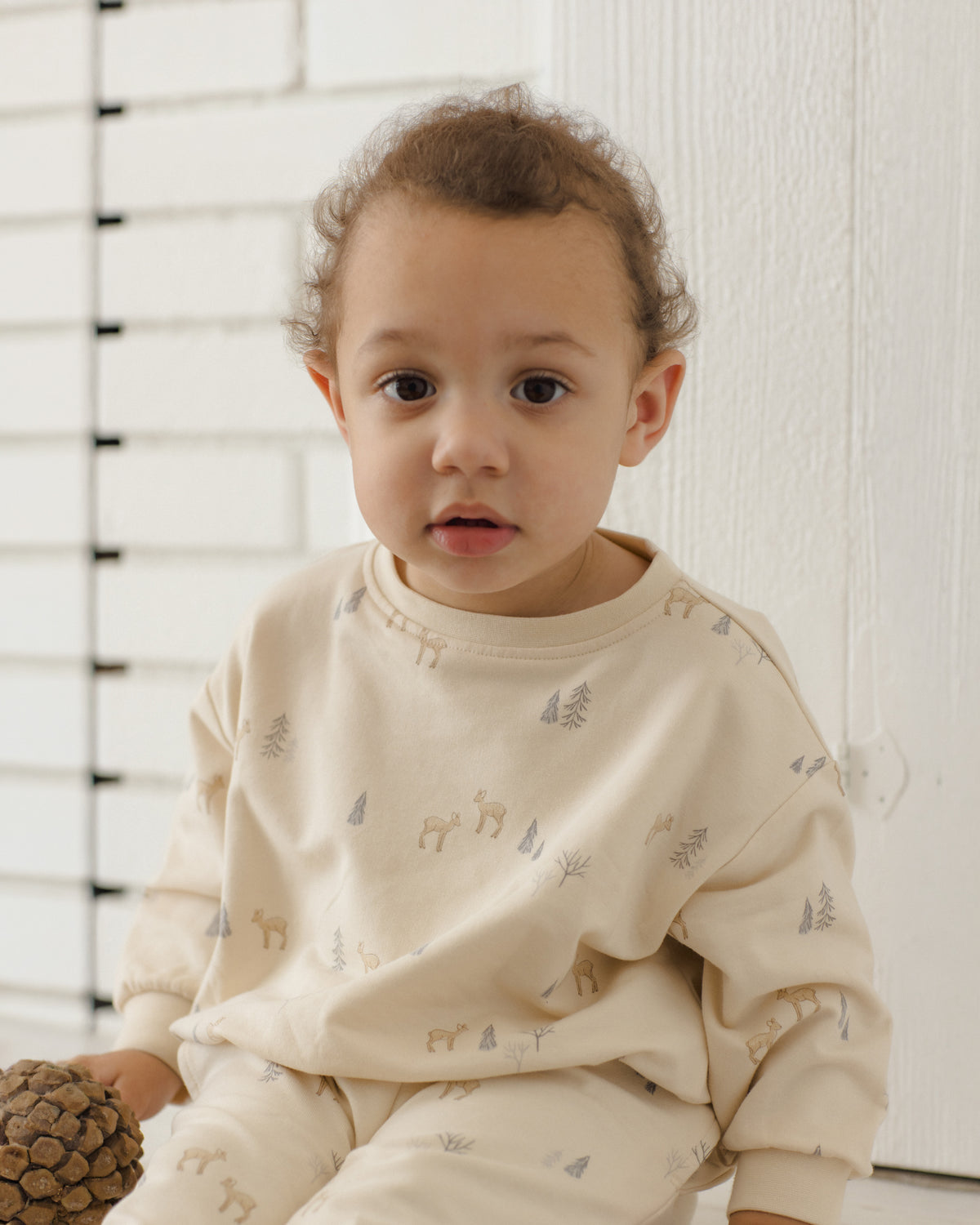 A toddler wearing a cream-colored long-sleeve pajama set with a subtle deer and tree print, sitting in front of a white paneled wall.