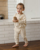 A toddler stands barefoot indoors in front of a white brick fireplace, wearing a cream-colored long-sleeve sweatshirt and matching sweatpants with a subtle allover animal print. The child is holding a small pinecone.