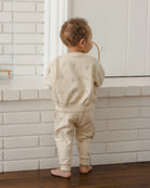 A toddler standing barefoot in front of a white brick wall, wearing a light beige long-sleeve sweatshirt and matching pants with a small repeating animal print.