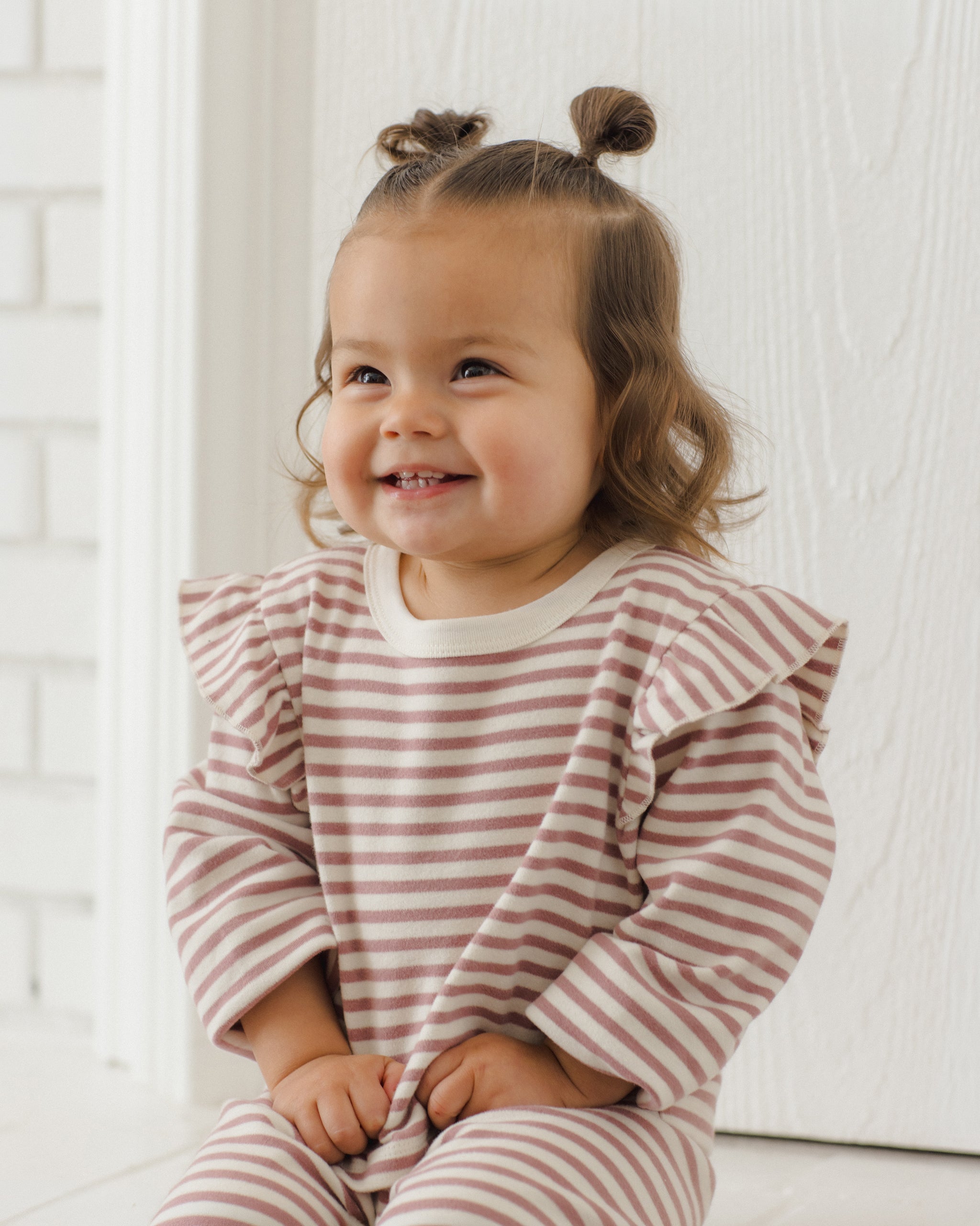 A smiling toddler with two buns sits wearing a long-sleeve ruffled shirt and matching striped pants in a soft pink and white pattern, set against a bright white interior background.