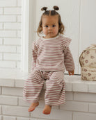A toddler girl with two small buns sits barefoot on a white brick ledge, wearing a matching long-sleeve top and wide-leg pants set in a dusty pink and cream stripe pattern. A floral-patterned bag rests on the right side of the ledge.
