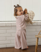 A toddler stands barefoot indoors against a white paneled wall, wearing a matching two-piece pajama set with thin brown stripes on a warm off-white background. The set includes a long-sleeve top and wide-leg pants.