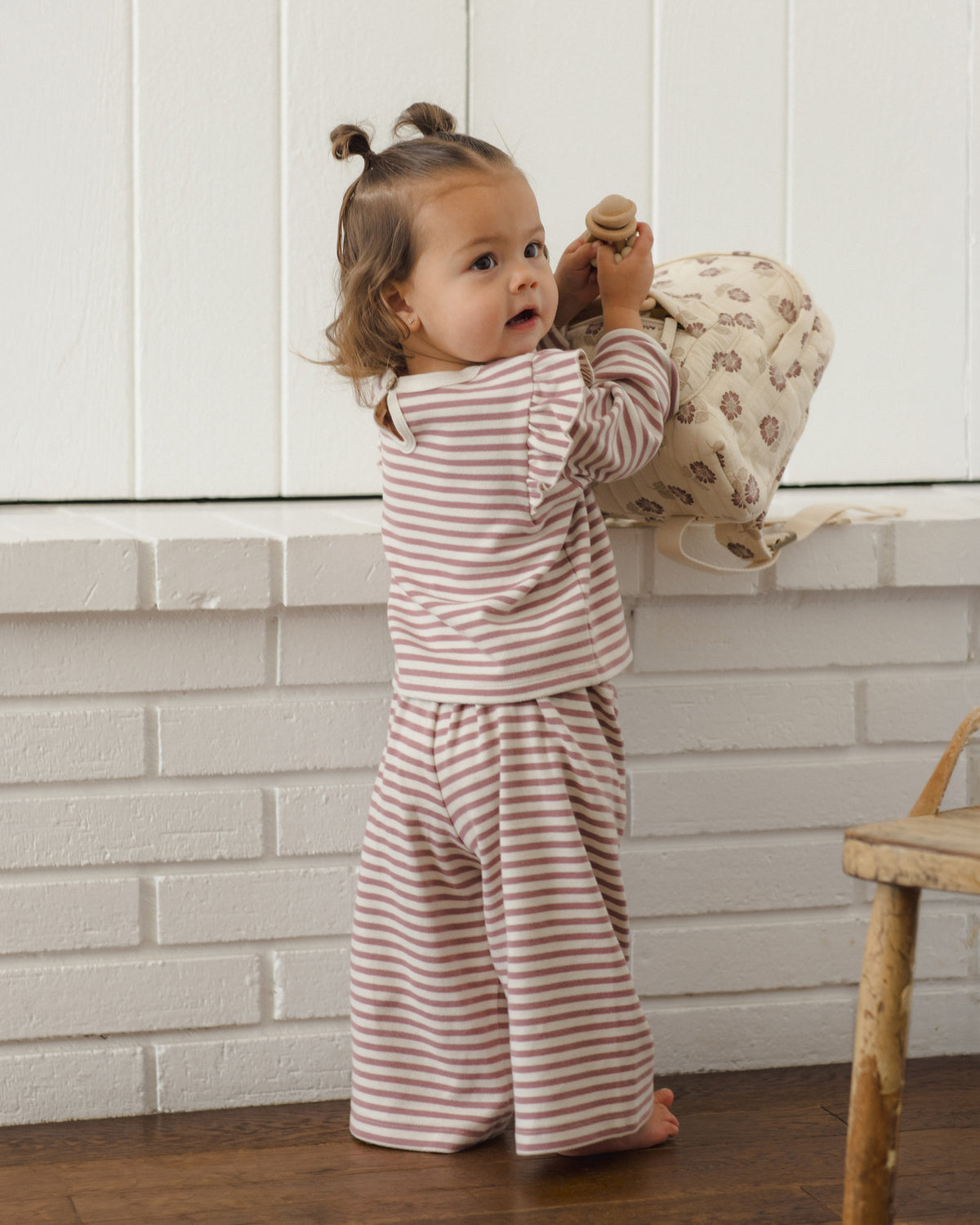 A toddler stands barefoot indoors against a white paneled wall, wearing a matching two-piece pajama set with thin brown stripes on a warm off-white background. The set includes a long-sleeve top and wide-leg pants.