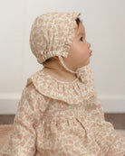 A baby wearing a matching beige and dusty pink floral bonnet and long-sleeve dress, shown in profile against a soft off-white background.