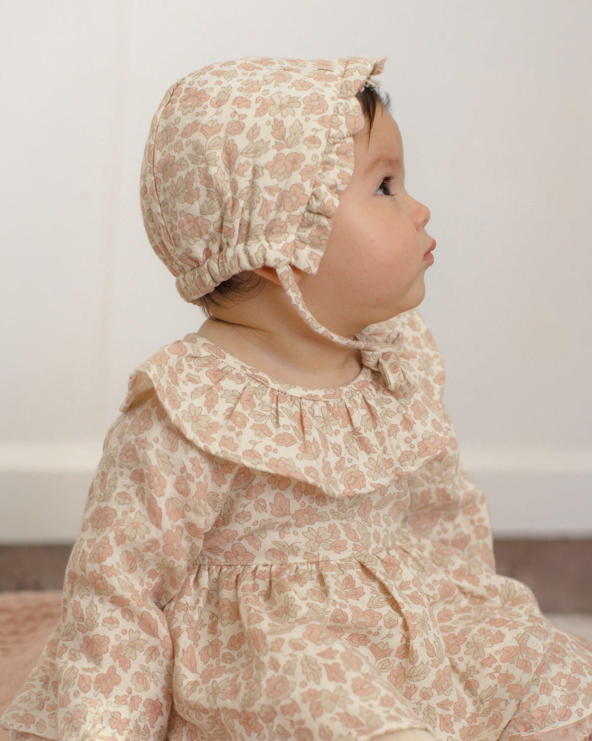 A baby wearing a matching beige and dusty pink floral bonnet and long-sleeve dress, shown in profile against a soft off-white background.