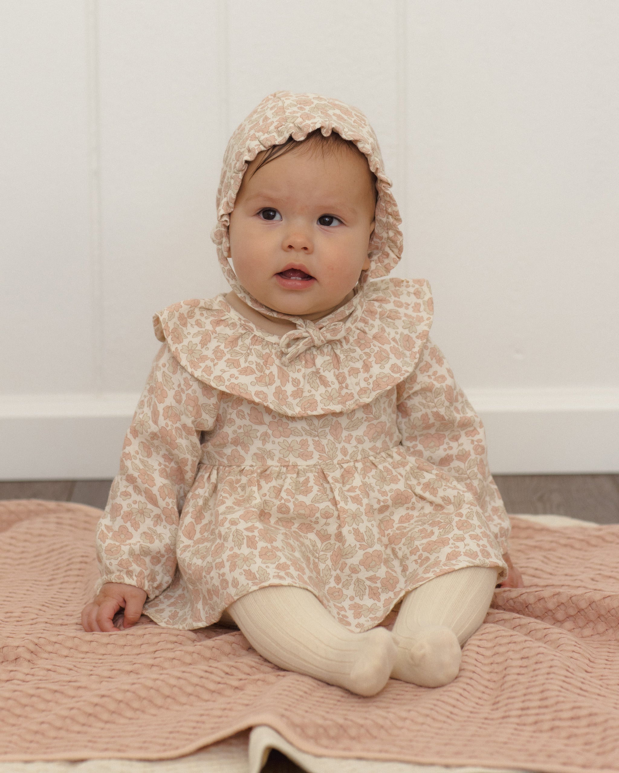 A baby wearing a beige and pink patterned dress with a matching bonnet, sitting on a pink blanket against a white paneled wall.