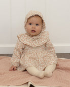 A baby wearing a beige and pink patterned dress with a matching bonnet, sitting on a pink blanket against a white paneled wall.