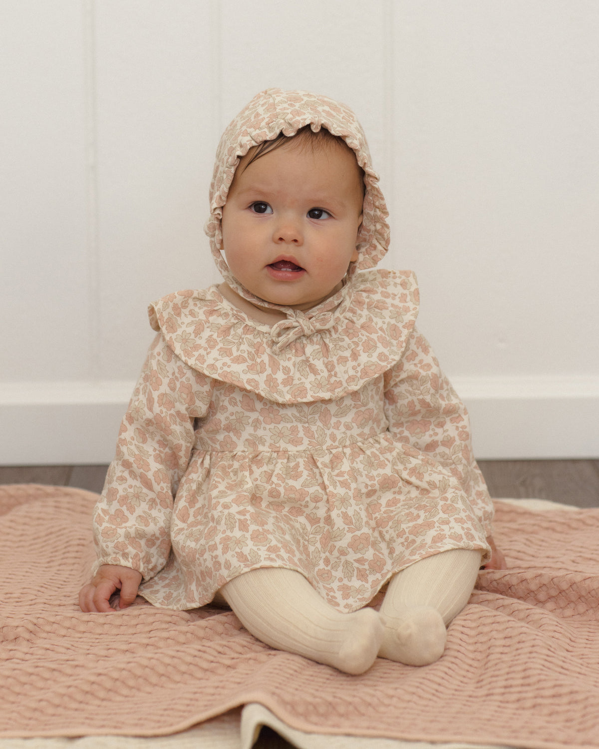 A baby wearing a beige and pink patterned dress with a matching bonnet, sitting on a pink blanket against a white paneled wall.