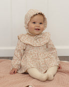 A baby wearing a light beige and pink floral print dress with a large ruffled collar, matching bonnet, and cream tights, sitting on a pink quilted blanket against a white paneled wall.