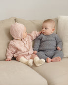 Two babies sitting on a beige sofa wearing matching knitted outfits. The baby on the left wears a light pink textured cardigan and bonnet with cream socks, and the baby on the right wears a dark gray textured cardigan and bonnet with cream socks.