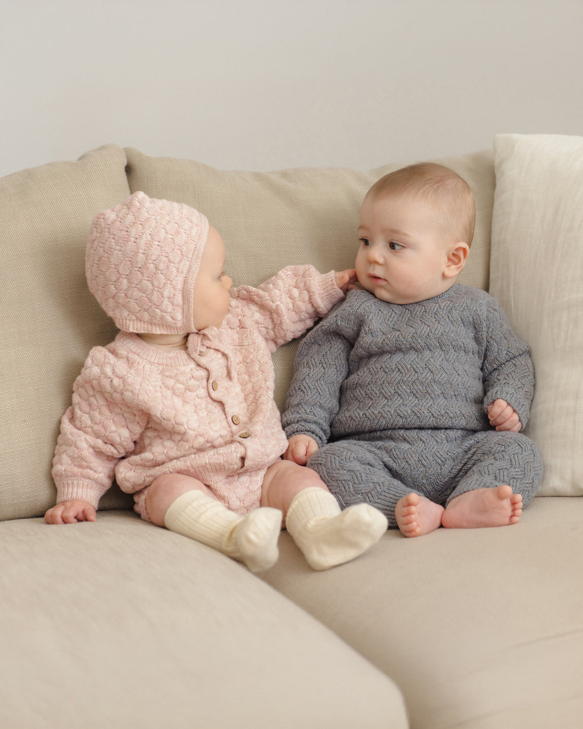 Two babies sitting on a beige sofa wearing matching knitted outfits. The baby on the left wears a light pink textured cardigan and bonnet with cream socks, and the baby on the right wears a dark gray textured cardigan and bonnet with cream socks.
