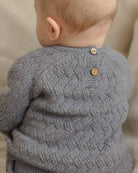 Close-up back view of a baby wearing a gray textured knit sweater with two wooden buttons at the neckline, set against a soft beige background.
