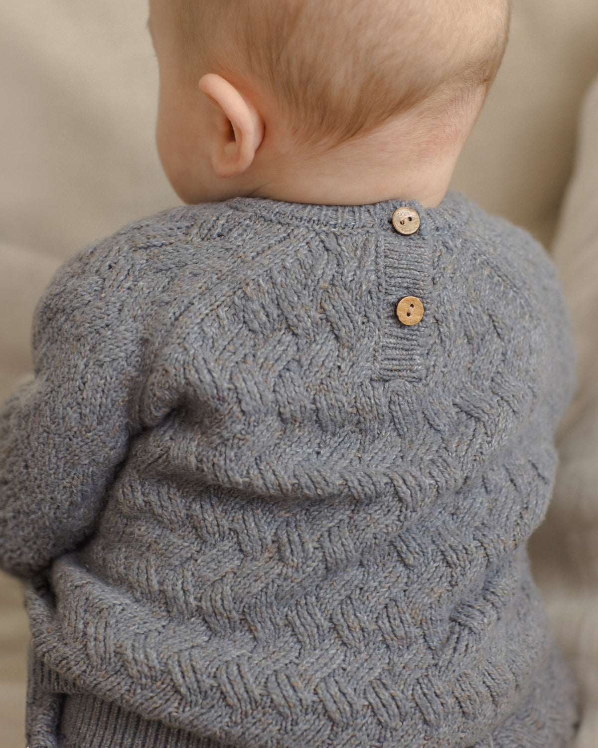 Close-up back view of a baby wearing a gray textured knit sweater with two wooden buttons at the neckline, set against a soft beige background.
