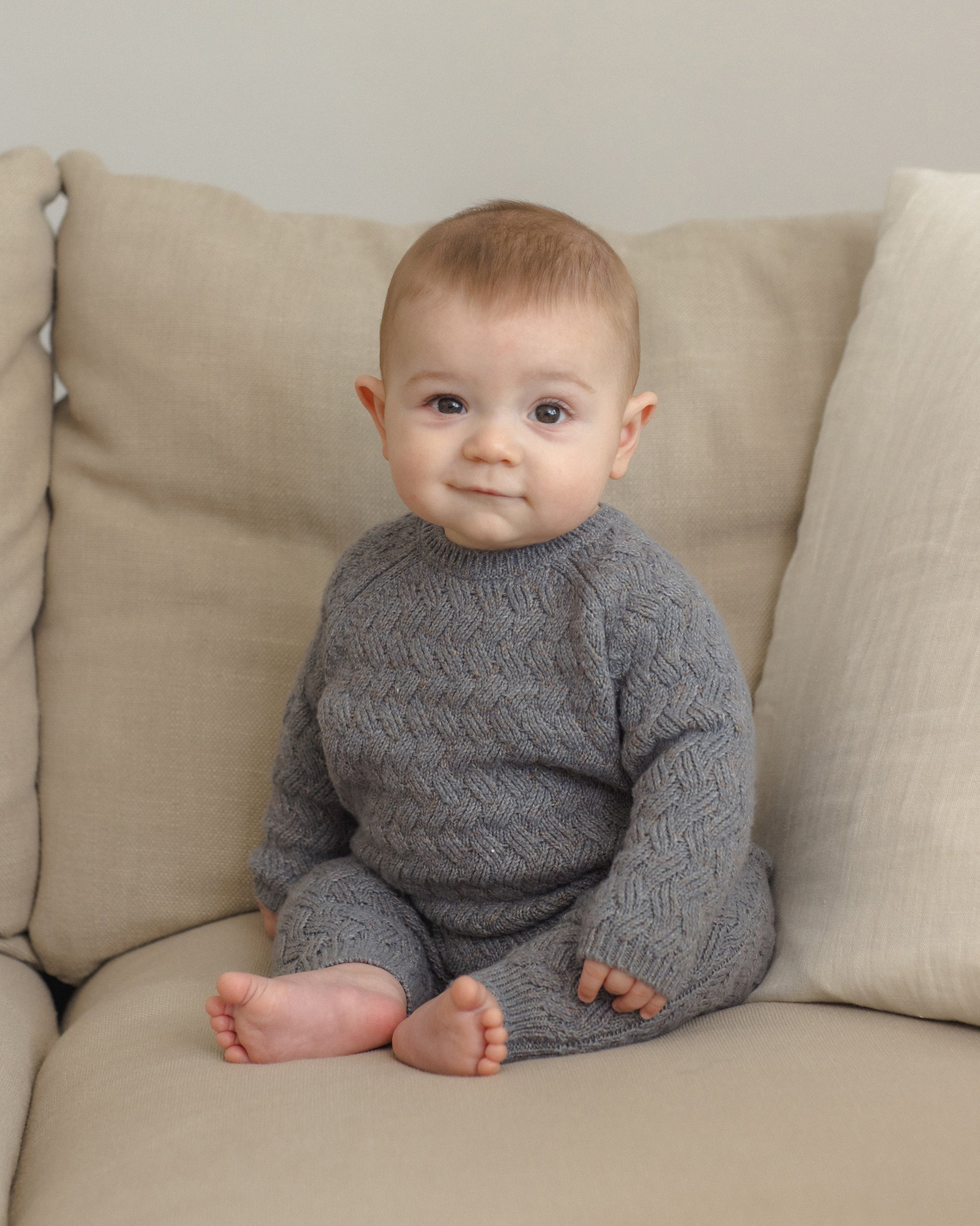 A baby sitting on a beige sofa wearing a gray cable-knit long-sleeve romper.