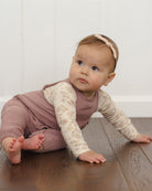 A baby crawling on a dark wood floor wearing a mauve-brown sleeveless jumpsuit over a cream floral long-sleeve shirt, with a matching pink headband.