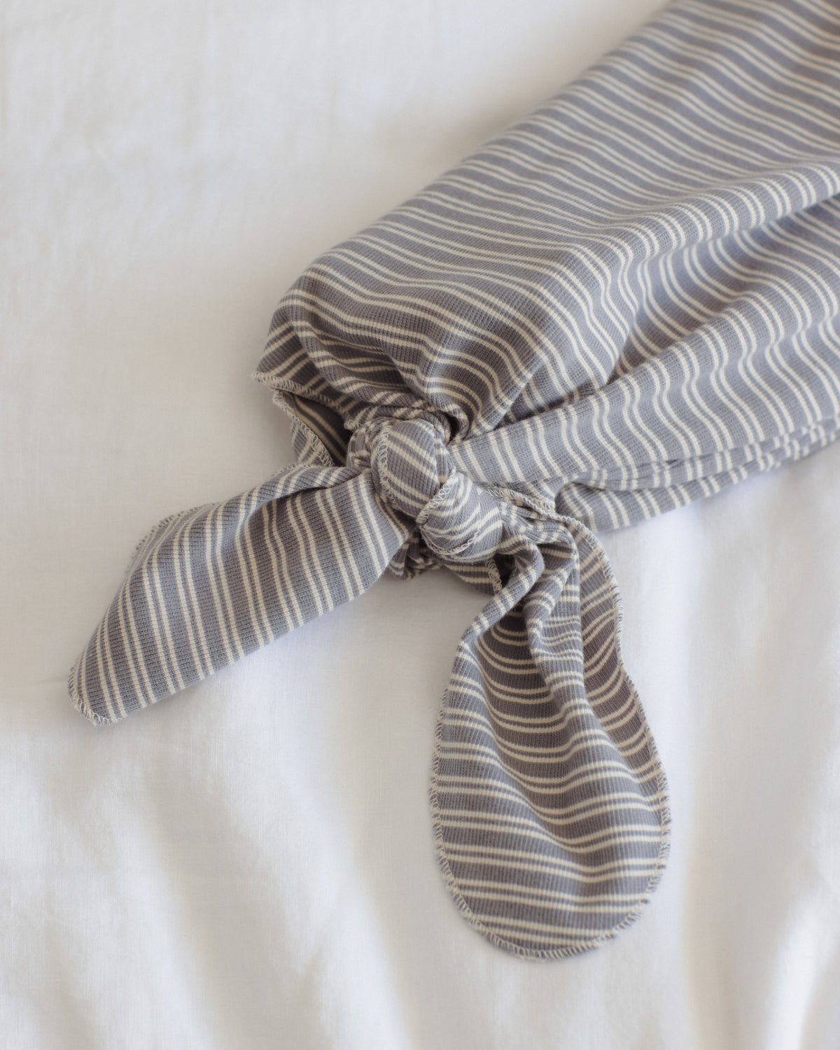 A gray and white striped fabric headband or hair tie, knotted at the front, displayed on a soft white fabric background.
