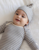 A baby lying on a white blanket, wearing a gray and white striped long-sleeve bodysuit and a matching striped beanie with a small bow on the tip.
