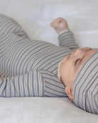 A baby lying on a white blanket, wearing a gray and white striped long-sleeve footless sleeper and a matching striped bonnet.